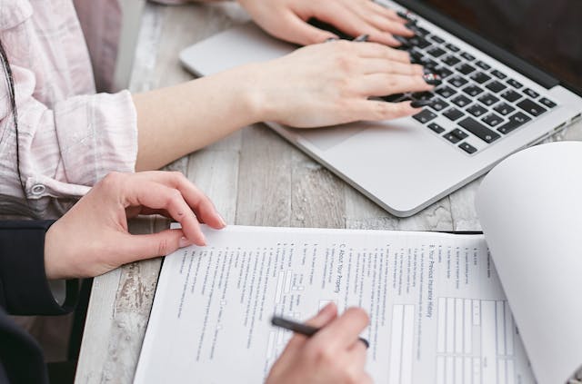 Person writing on paper with another person using a laptop next to them